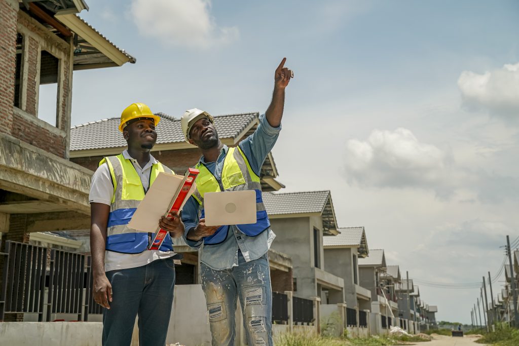 Two construction workers, wearing helmets and reflective vests, evaluate building plans while observing ongoing work in a residential area. The sky is bright and clear, showcasing a productive day.
